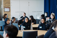 Students sit at round tables in a classroom, smiling and engaged in discussion during a collaborative learning or workshop session.